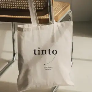 Tote bag de lona natural con el logo tinto y la frase café negro sin leche, colgada de una silla de esterilla