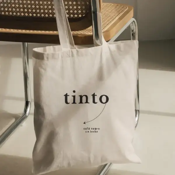Tote bag de lona natural con el logo tinto y la frase café negro sin leche, colgada de una silla de esterilla